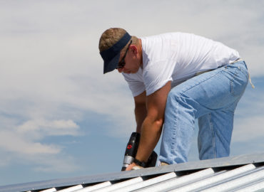 Construction worker uses a power drill to attach a cap the the top of a sheet metal roofing job with screws.